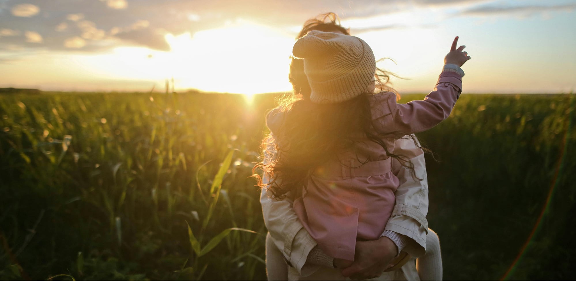 Adult holding child in a corn field at sunset.